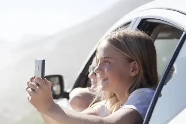 Smiling child enjoying a road trip while capturing moments on her smartphone.